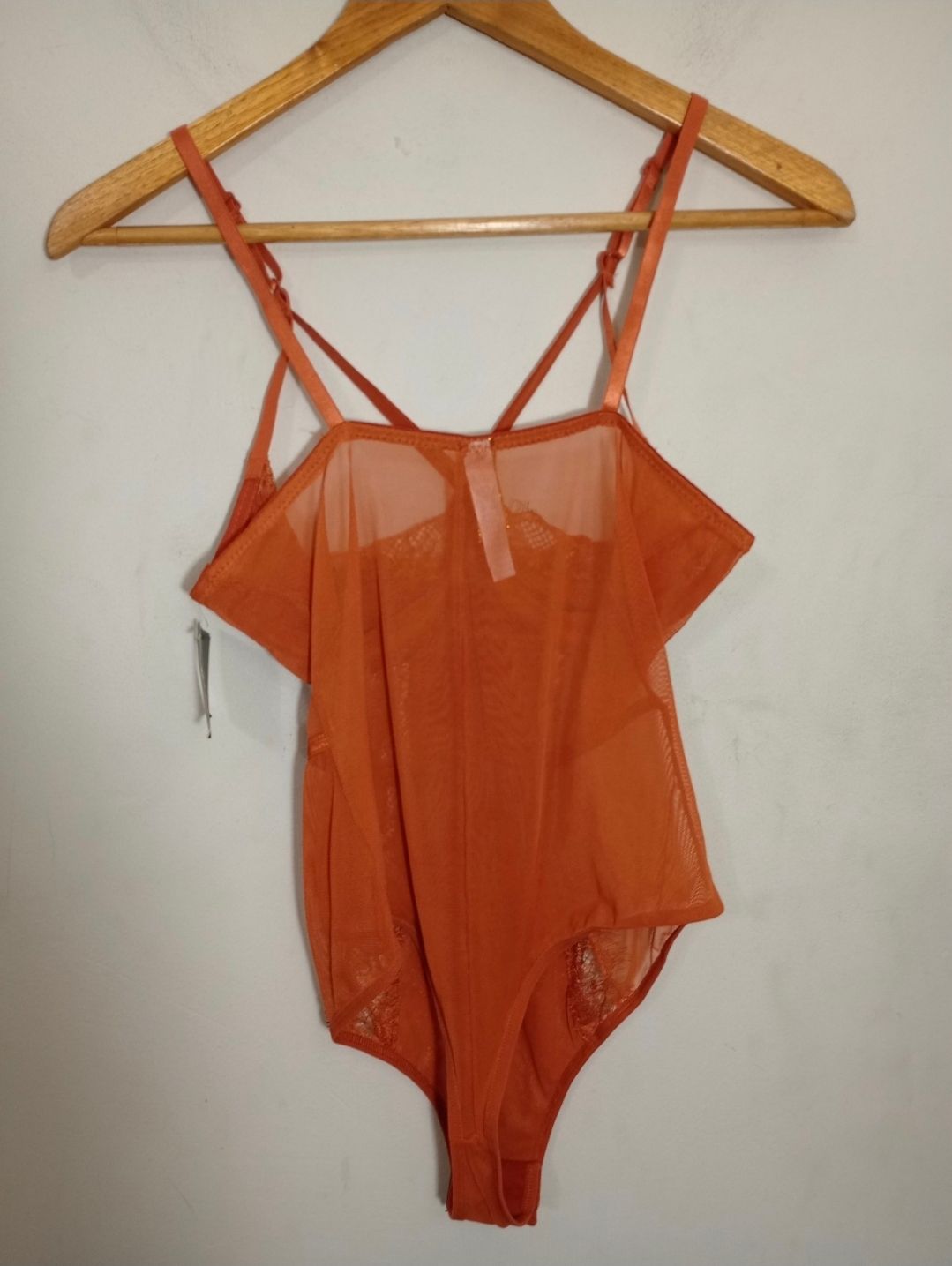 Orange mesh bodysuit with spaghetti straps and ruffle detail, hanging on wooden hanger with tags attached.