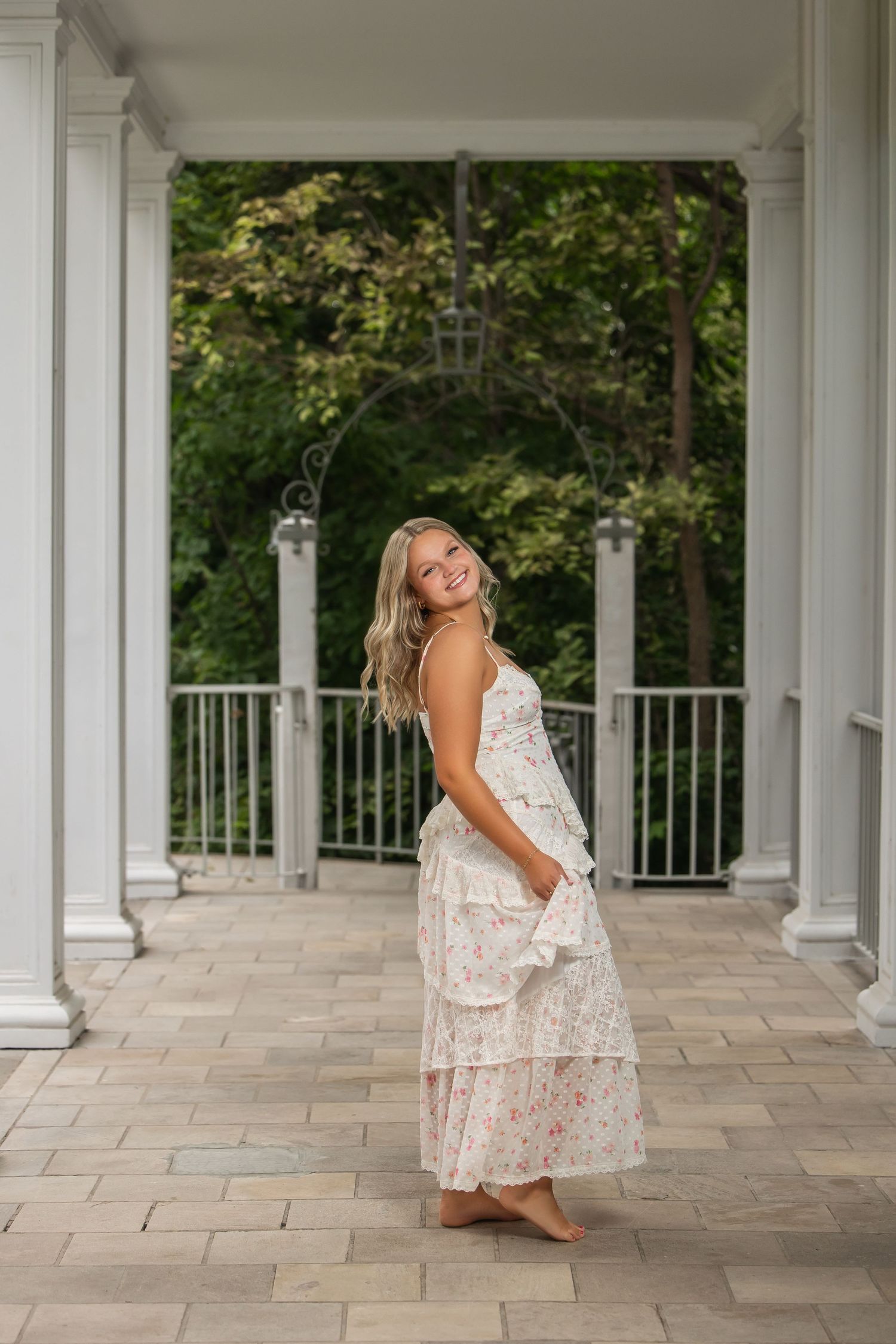 Young woman in floral maxi dress smiling on elegant porch with white columns and lush green trees.