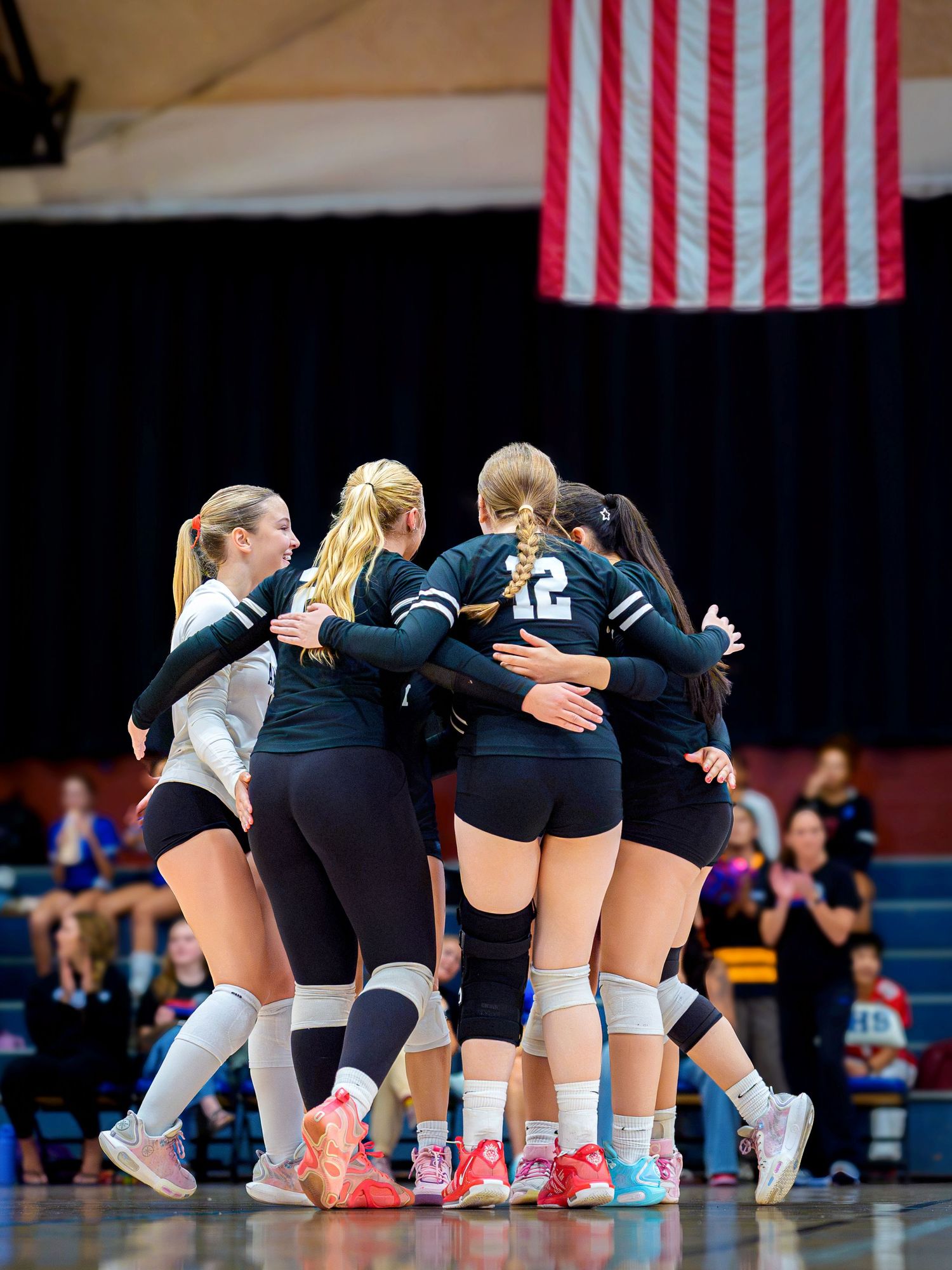Girls volleyball team huddles together in black uniforms on court with American flag in background.