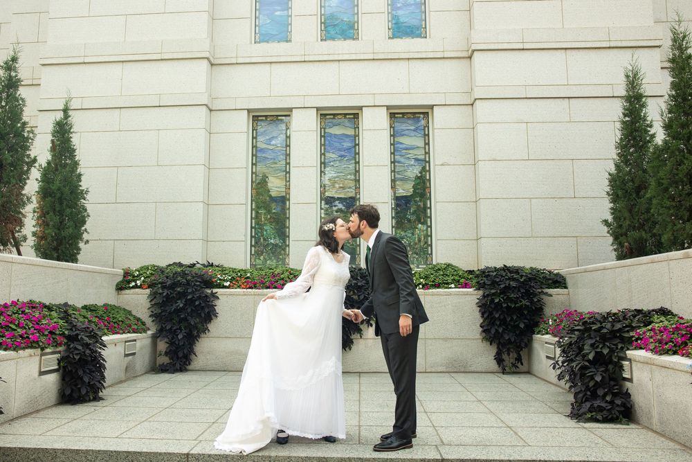 A bride and groom share a romantic kiss outside a grand white stone building surrounded by lush greenery and flowers.