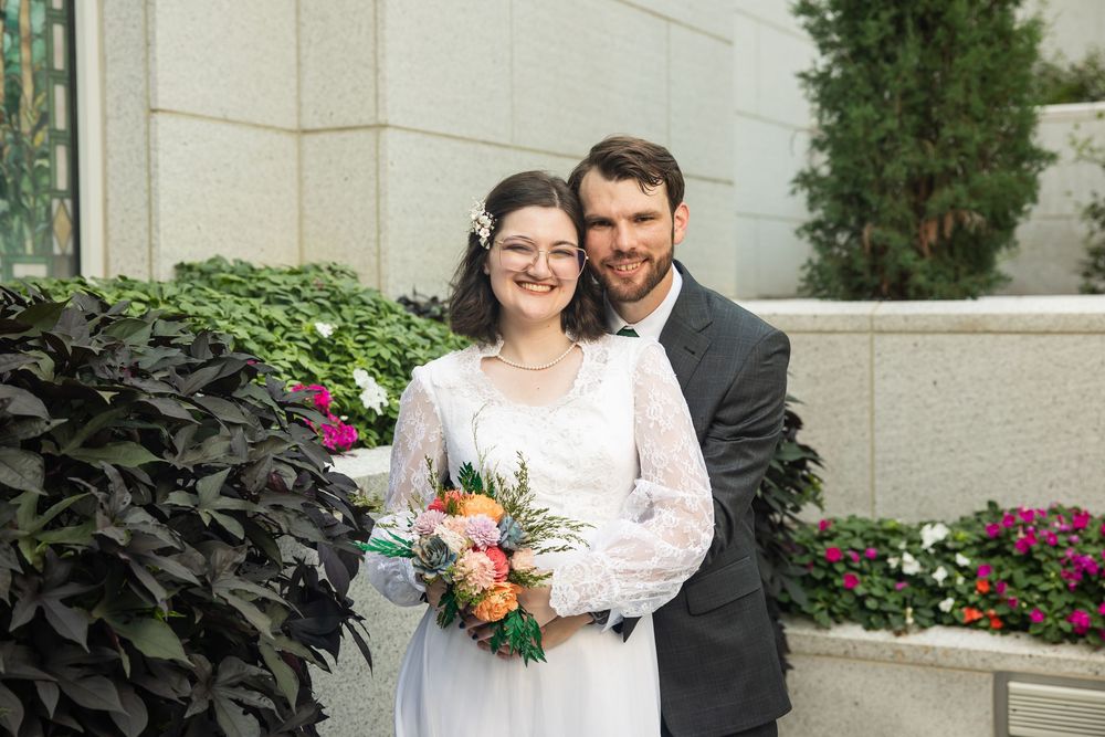 A smiling bride in a white lace gown holds a colorful bouquet beside her groom in a gray suit outdoors.