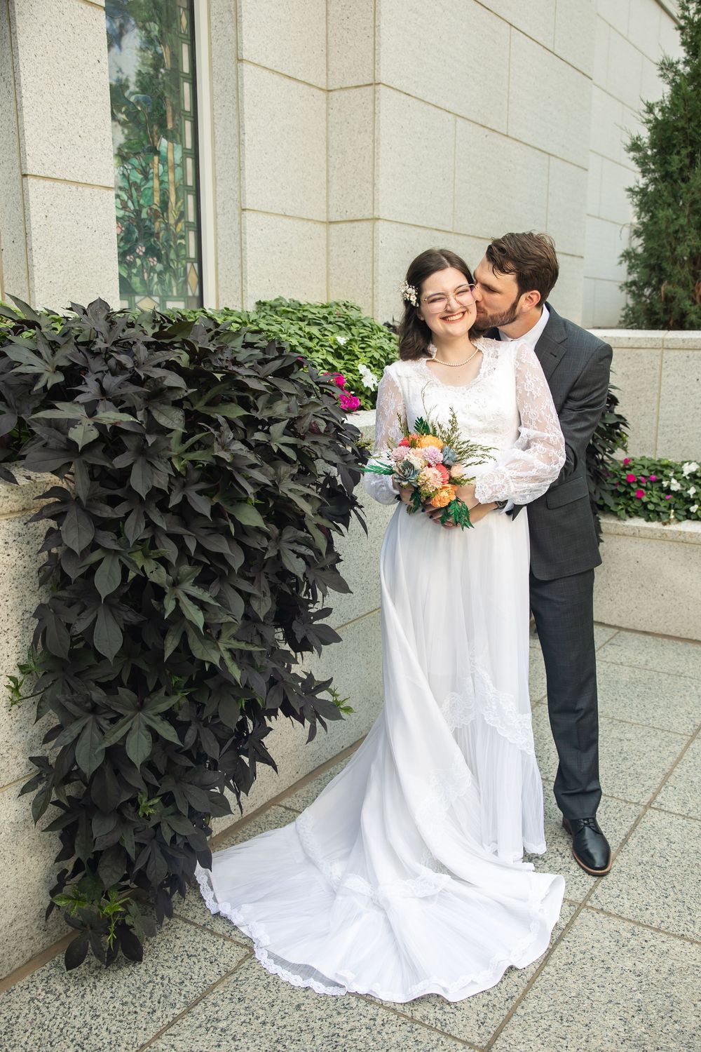 A bride in a white gown holds a colorful bouquet as the groom in a dark suit kisses her cheek outdoors.