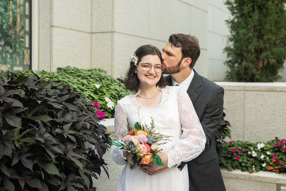 Bride in white lace gown holding colorful bouquet receives kiss from groom in dark suit outdoors near stone building.