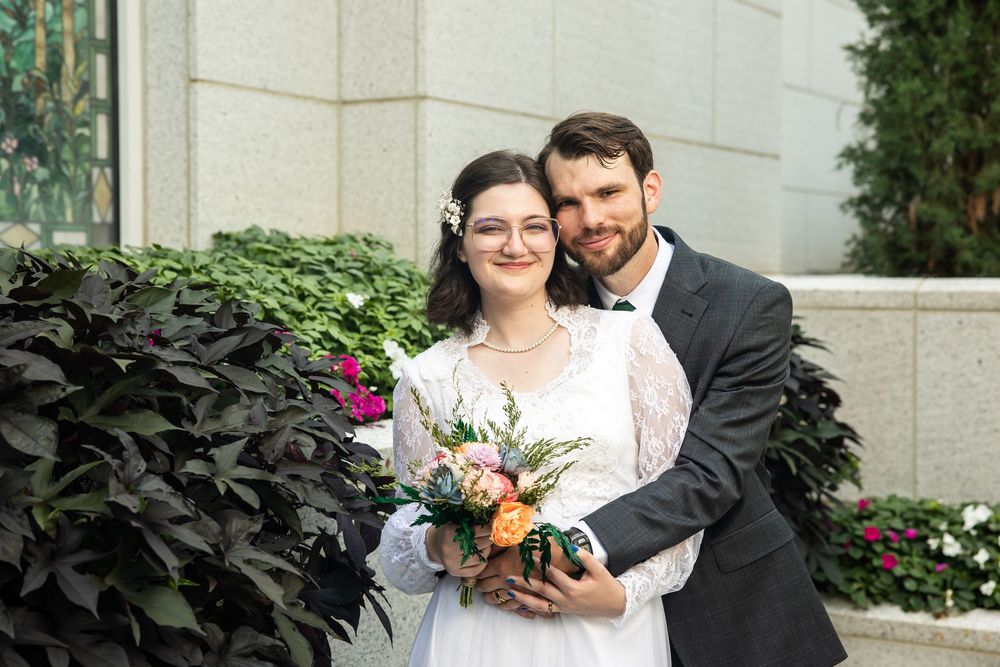Bride and groom pose together outdoors holding a colorful bouquet, with lush greenery and a stone building behind them.