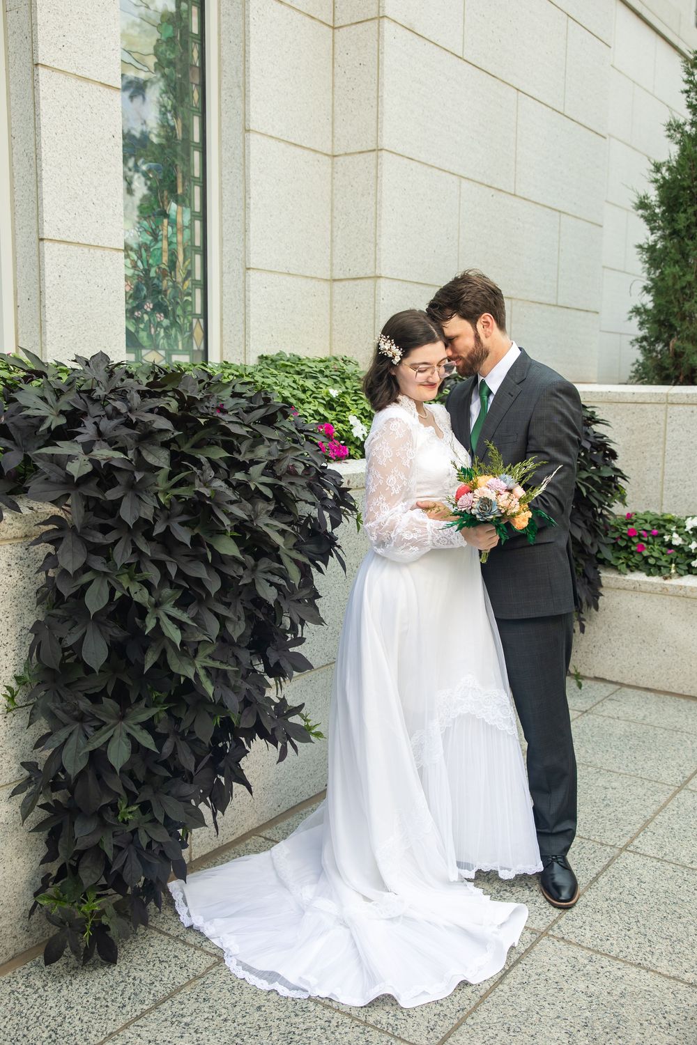 Bride in white lace gown and groom in gray suit share a tender moment outdoors, holding a colorful wedding bouquet.