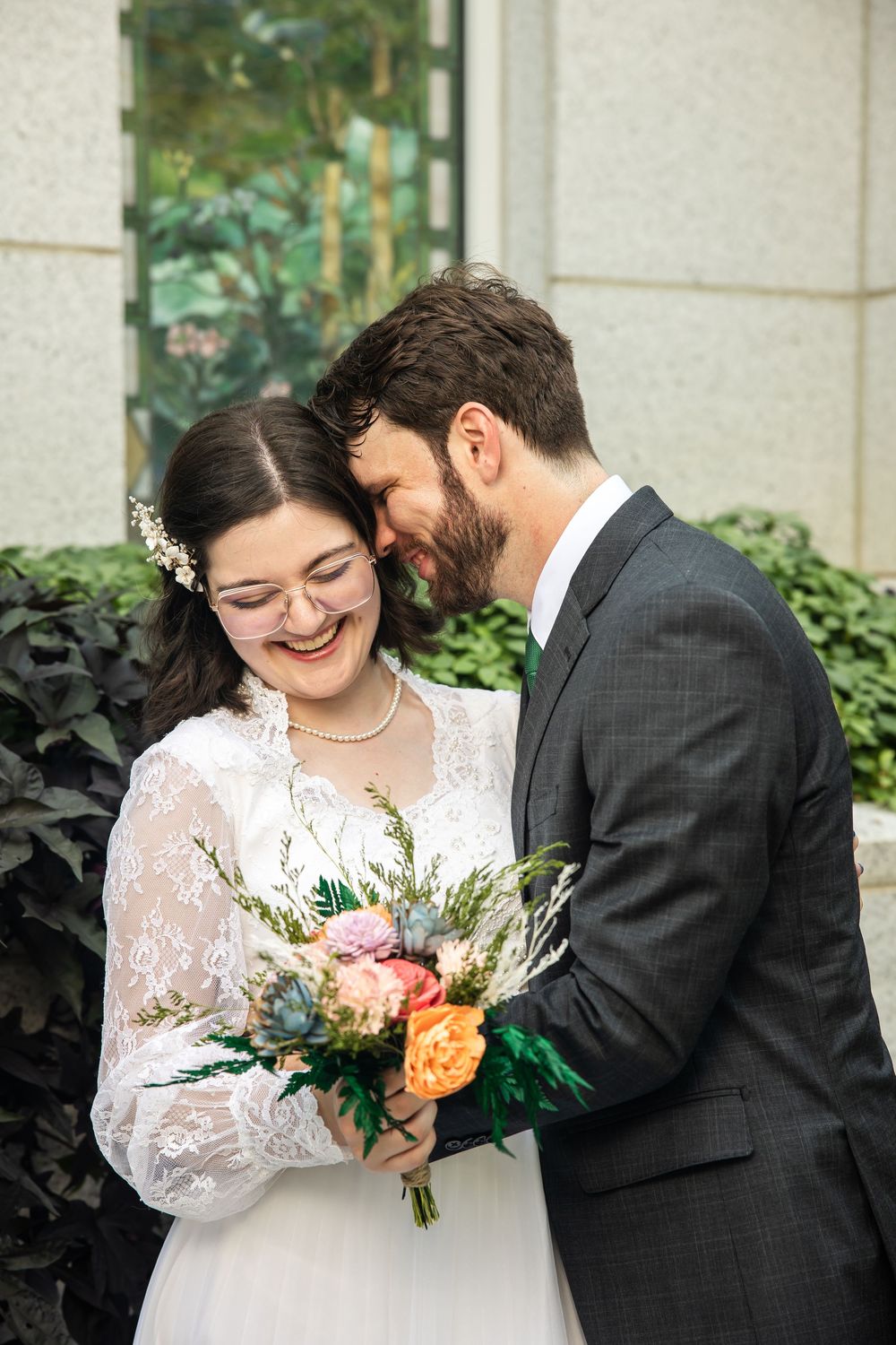 Bride and groom share a joyful moment, bride holding colorful bouquet in lace dress, groom in gray suit, outdoors.