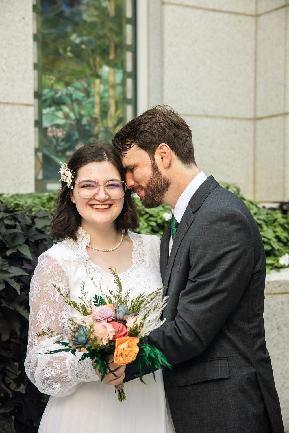 Smiling bride in lace dress holding colorful bouquet as groom in gray suit nuzzles her cheek outdoors.