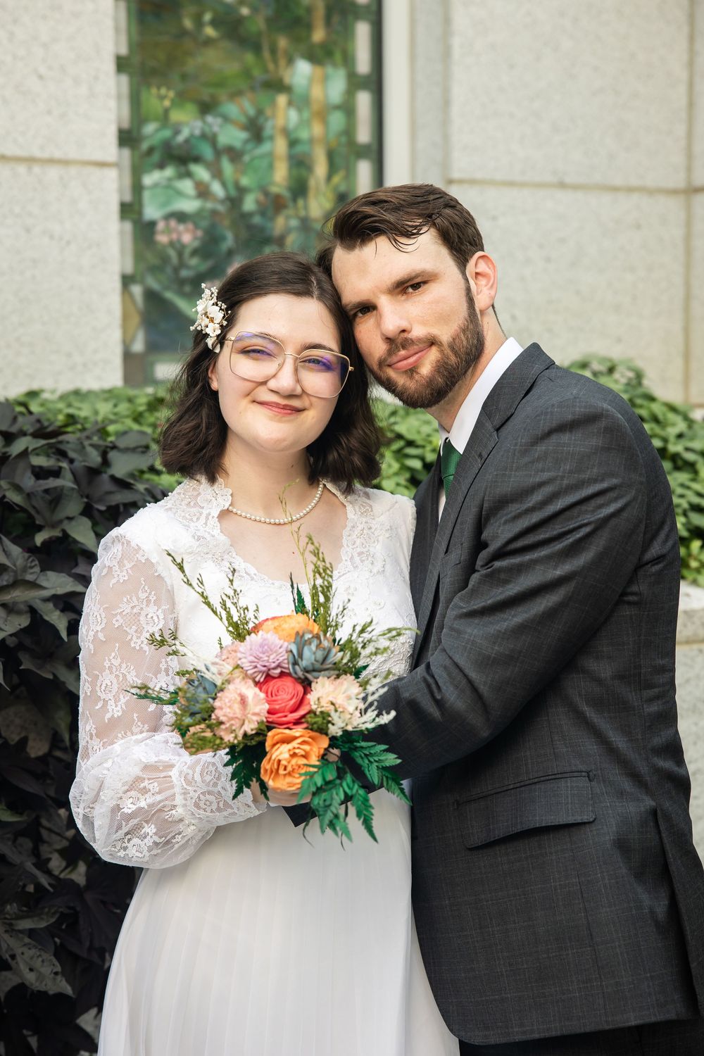 A bride in lace dress holding colorful bouquet poses with bearded groom in gray suit outdoors.
