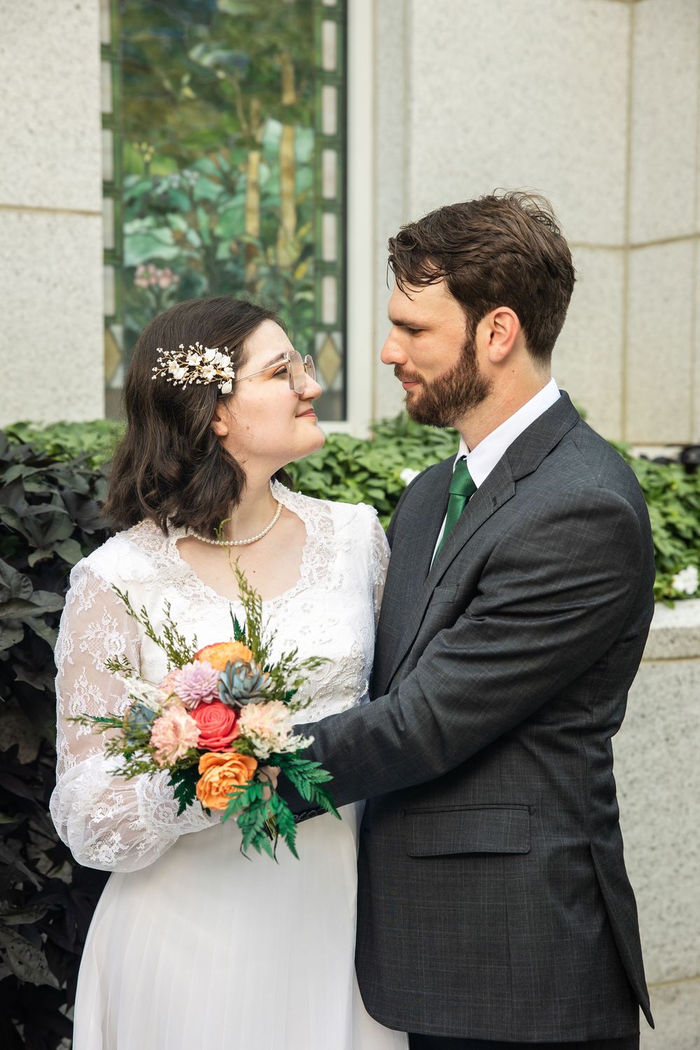 Bride and groom gazing at each other, bride holding colorful bouquet, groom in gray suit with green tie, outdoor wedding portrait.