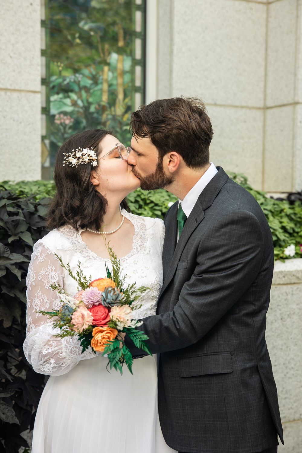 Bride and groom share a kiss outdoors, bride holding colorful bouquet in white lace dress, groom in gray suit with green tie.