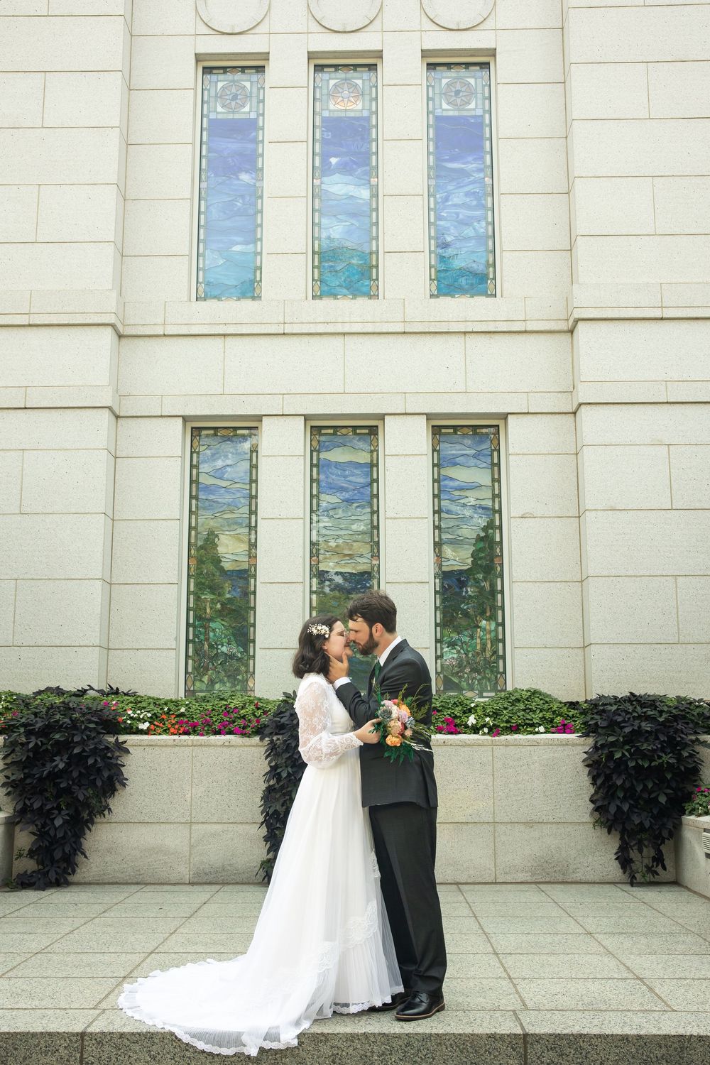 Bride and groom kissing in front of a white stone building with colorful stained glass windows on their wedding day.