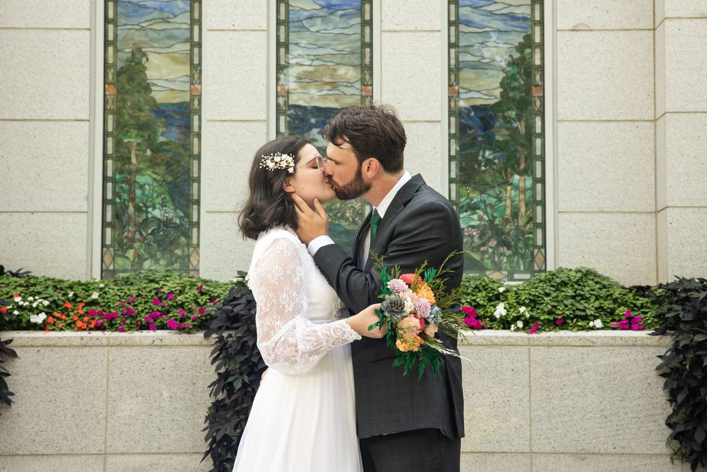 Newlywed couple sharing a romantic kiss outside a temple, bride holding colorful bouquet in white lace gown.