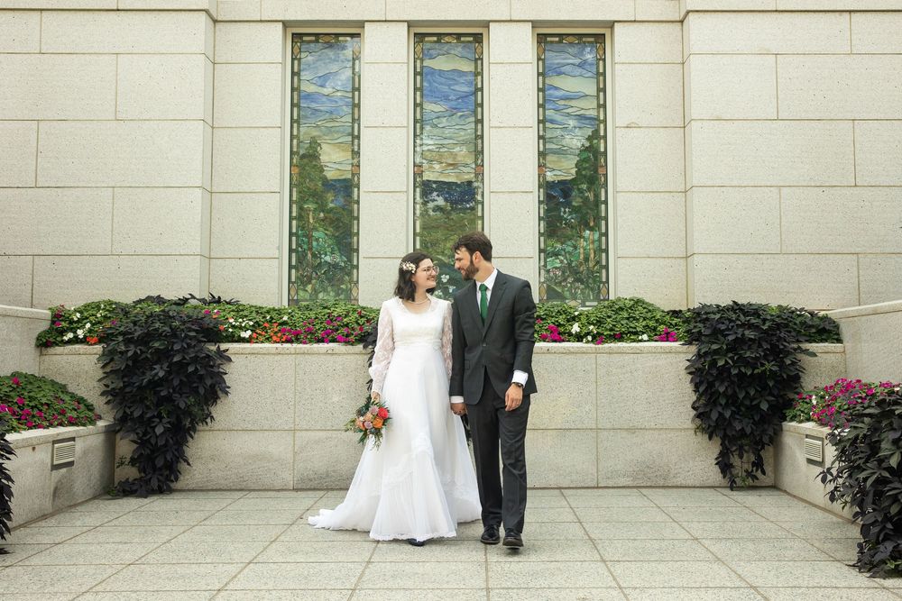 Bride and groom pose together outside a stone building with stained glass windows and flower beds on their wedding day.