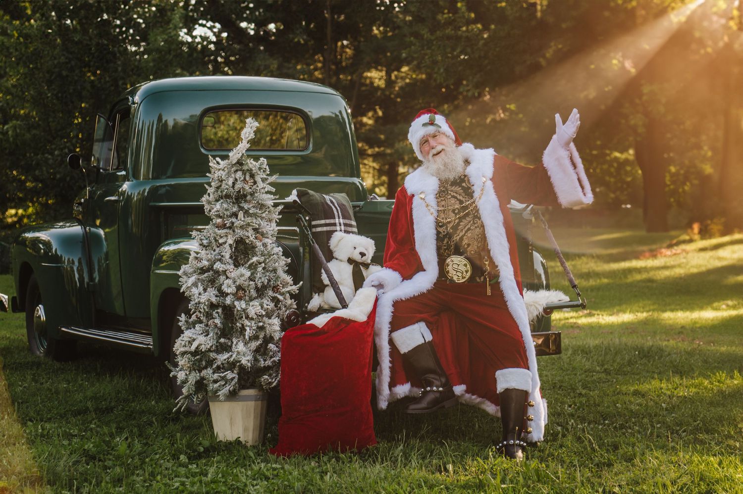 Santa Claus posing beside a vintage green truck with a flocked Christmas tree and gifts on a sunny day.