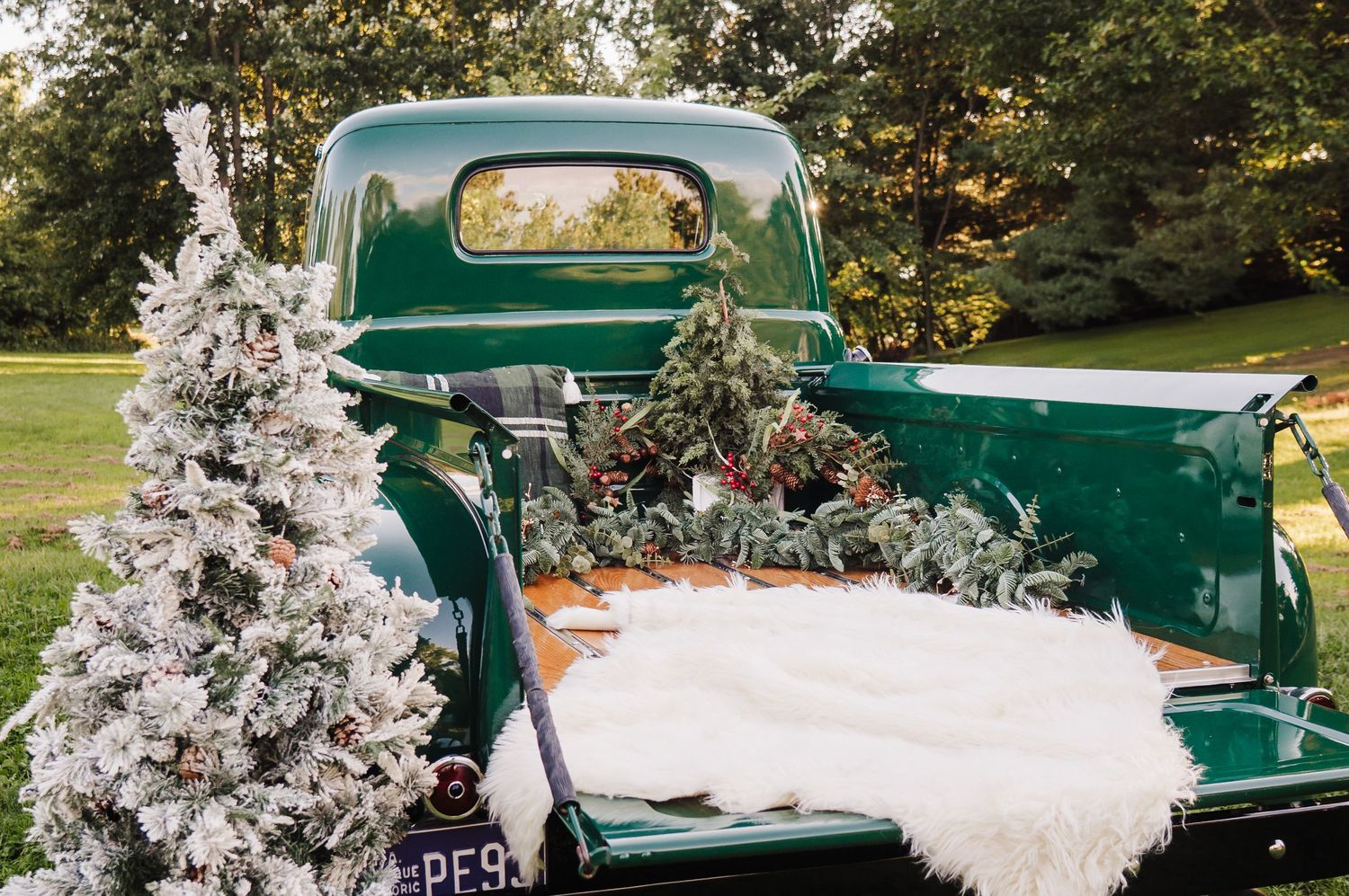 Vintage green pickup truck decorated with flocked Christmas tree and holiday greenery on a lush lawn.