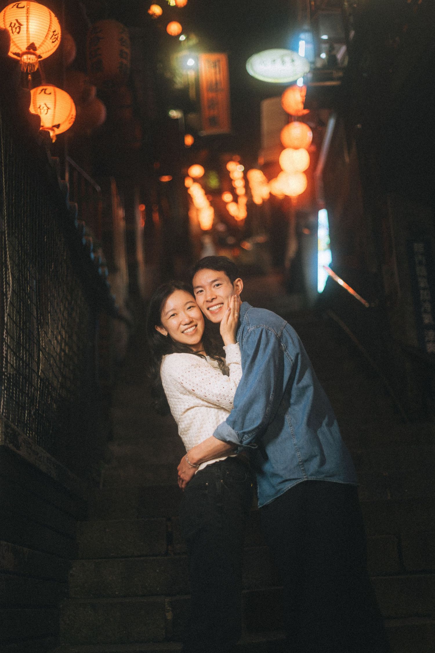 A couple embraces and smiles on a lantern-lit alley stairway at night in a traditional Asian town.