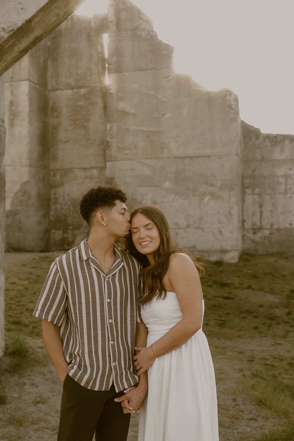 Couple sharing a tender kiss near rustic stone ruins during golden hour engagement session.