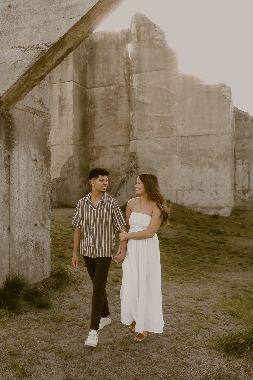 Couple walking hand in hand past concrete ruins, man in striped shirt smiling at woman in white dress.