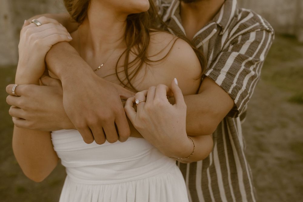 A couple embraces tenderly, the man wrapping his arms around the woman in a white dress, both wearing rings.