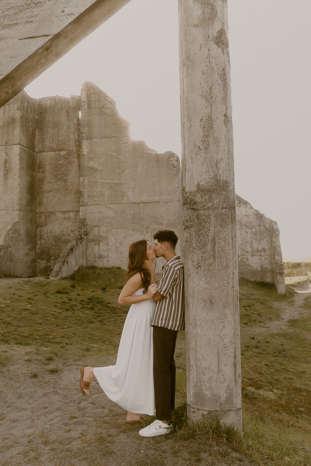 Couple sharing a romantic kiss beside a concrete pillar at ancient ruins, woman in white dress, man in striped shirt.