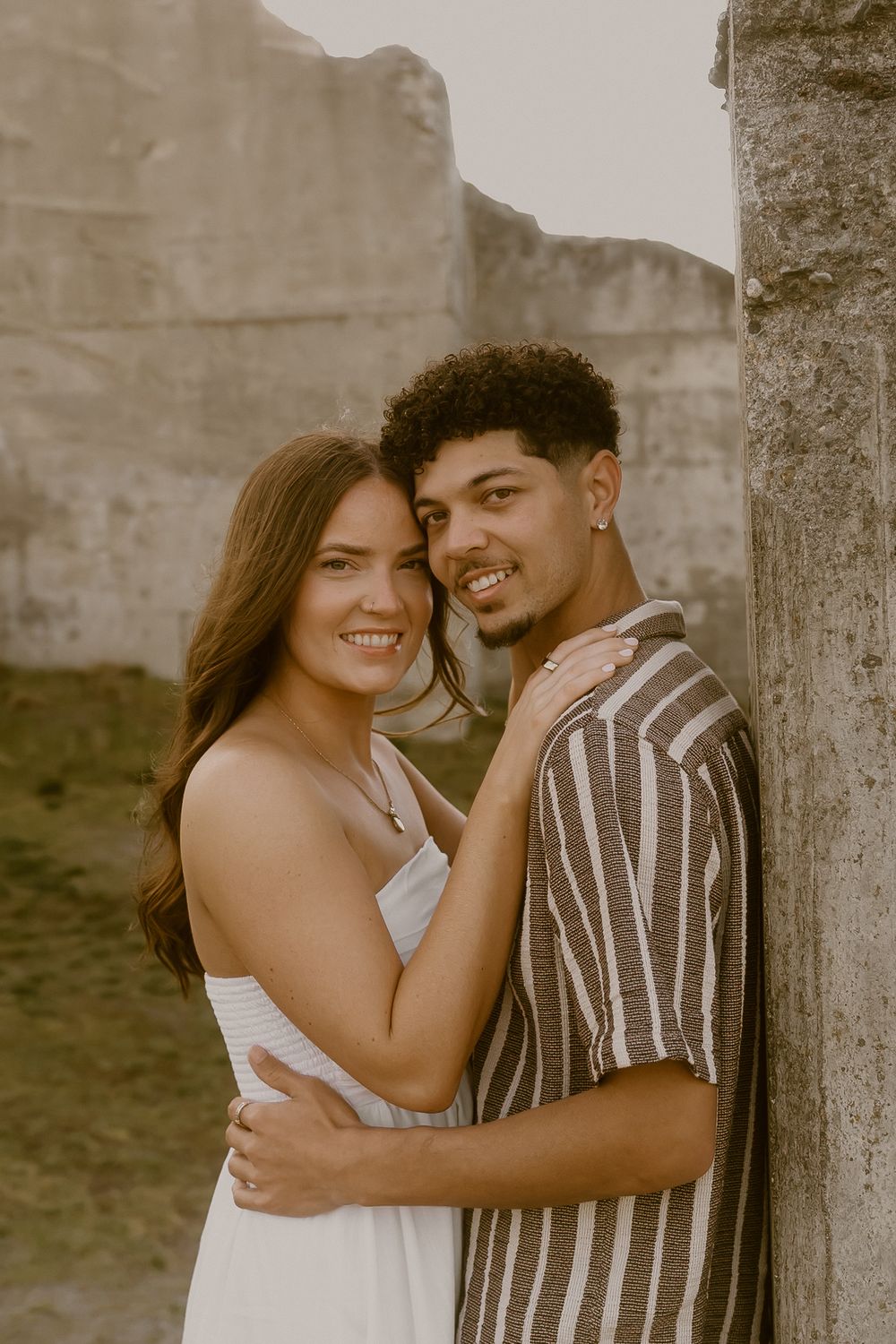 A smiling couple embraces near a stone column, the woman in a white dress and man in a striped shirt, in a warm outdoor setting.