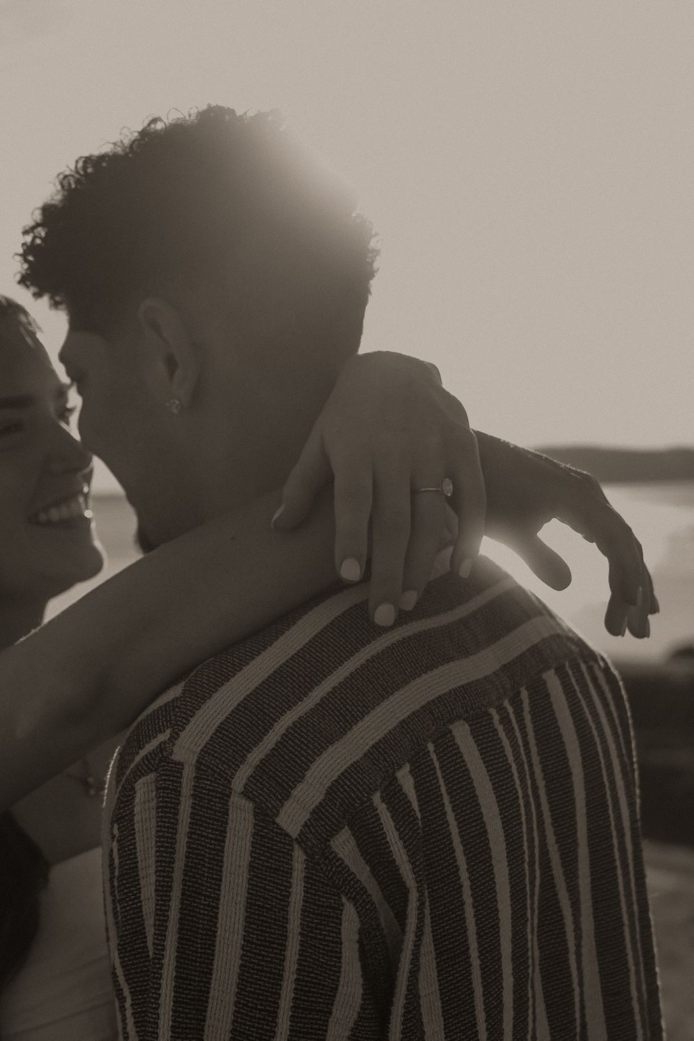 Black and white photo of a couple embracing at sunset, woman smiling with arm around mans shoulder wearing a ring.