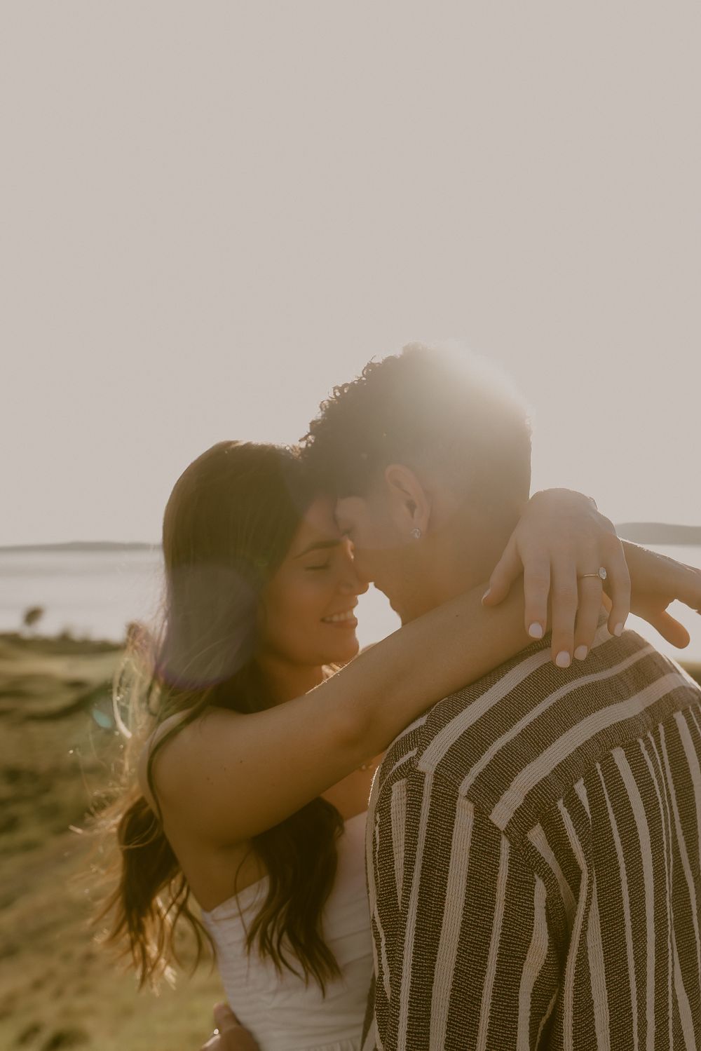 Couple sharing a romantic sunset moment outdoors, woman embracing man in striped shirt with golden backlight.
