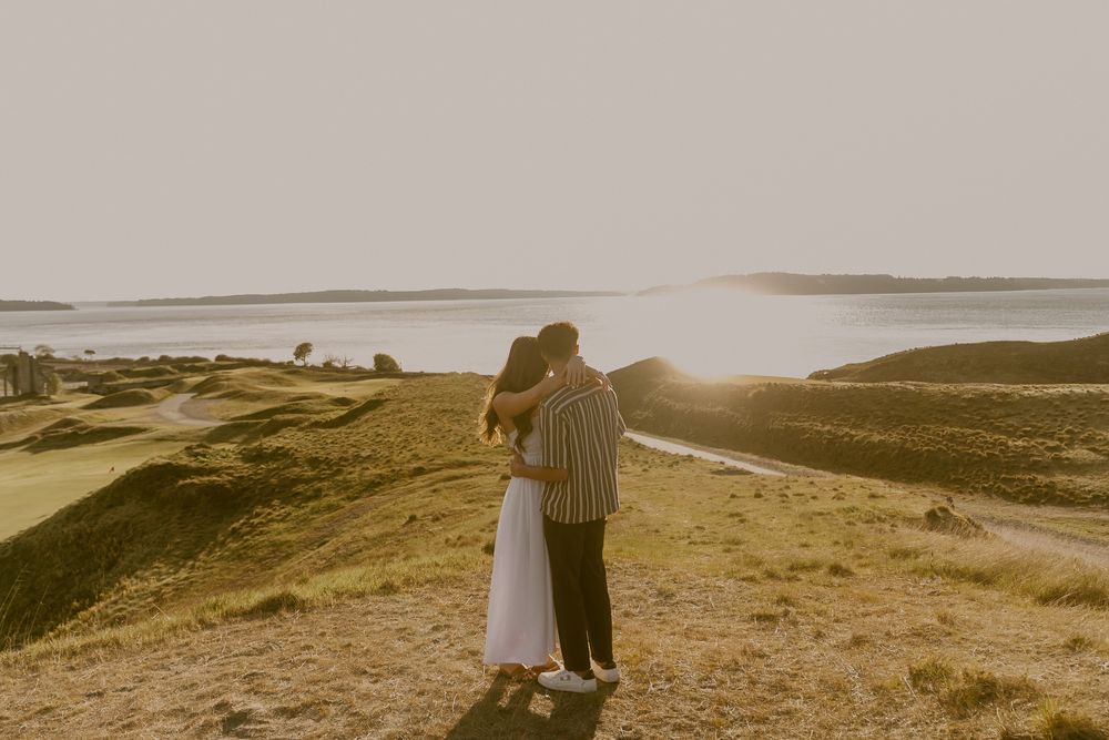 Couple embracing at golden hour on a coastal hillside with scenic water views in the background.