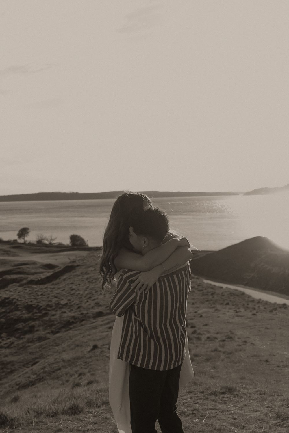 Couple embracing on a scenic hilltop overlooking a lake in a romantic black and white outdoor photo.