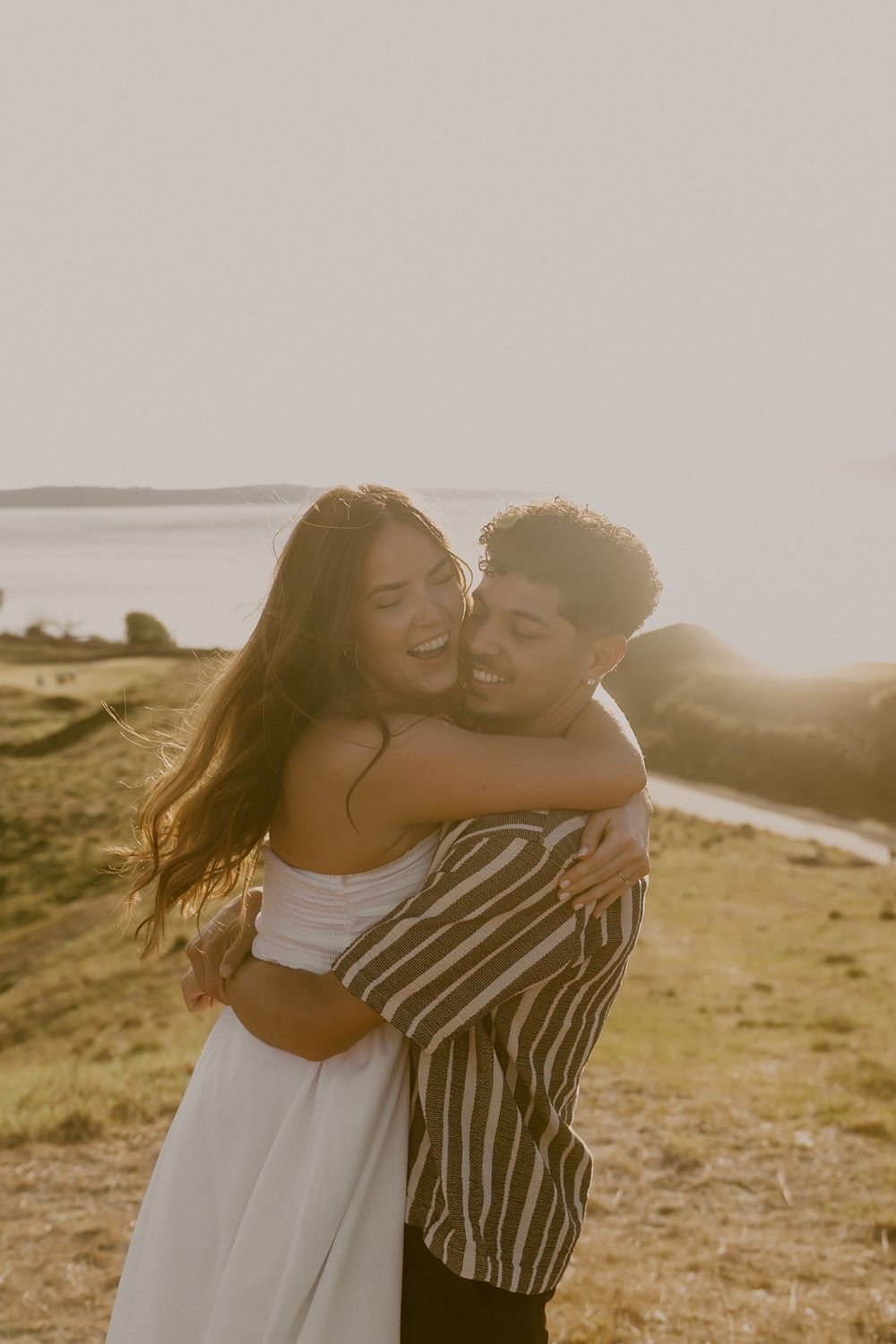 A couple embraces and laughs together at golden hour near a scenic waterfront, radiating joy and romance.