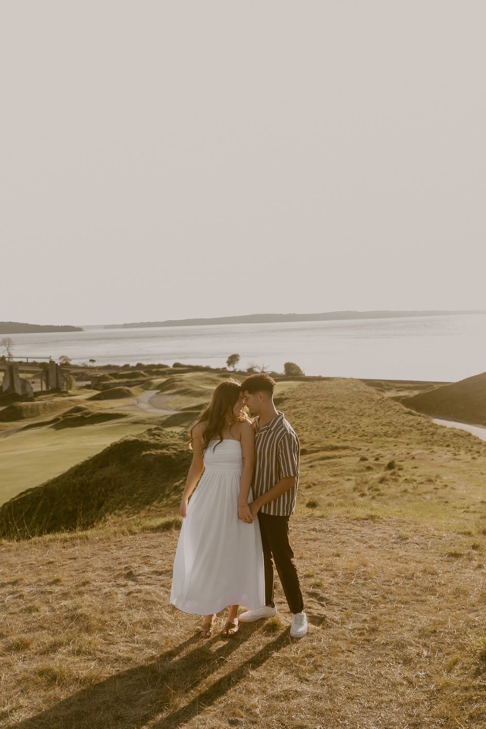 Romantic couple embracing on scenic coastal hillside during golden sunset engagement shoot.
