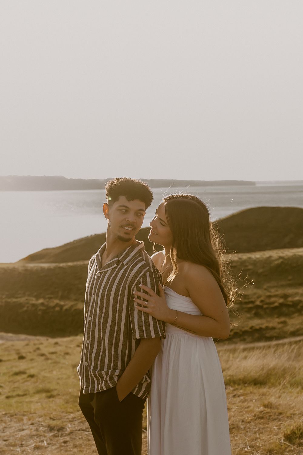 A couple embraces at golden hour near a coastal landscape, the man in a striped shirt and woman in a white dress.