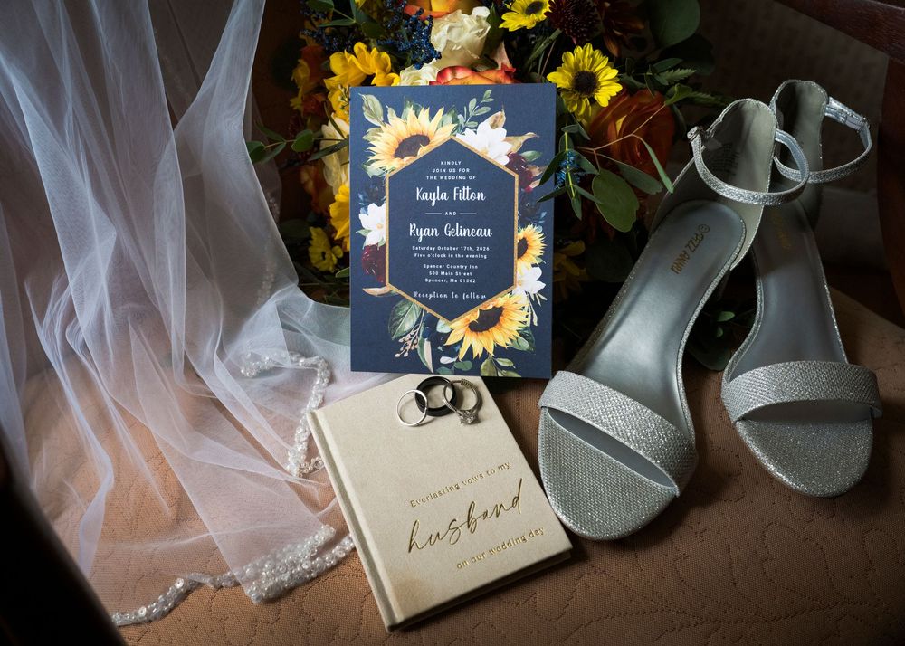 Sunflower wedding invitation, silver glitter heels, bridal veil, wedding rings and a husband keepsake book beside a floral bouquet.