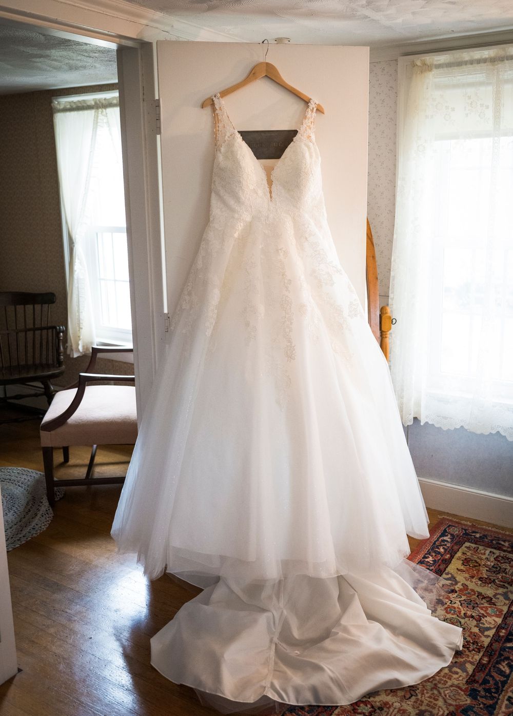 White lace wedding gown with a full tulle skirt and long train hanging on a wooden hanger in a bright room with a vintage rug.