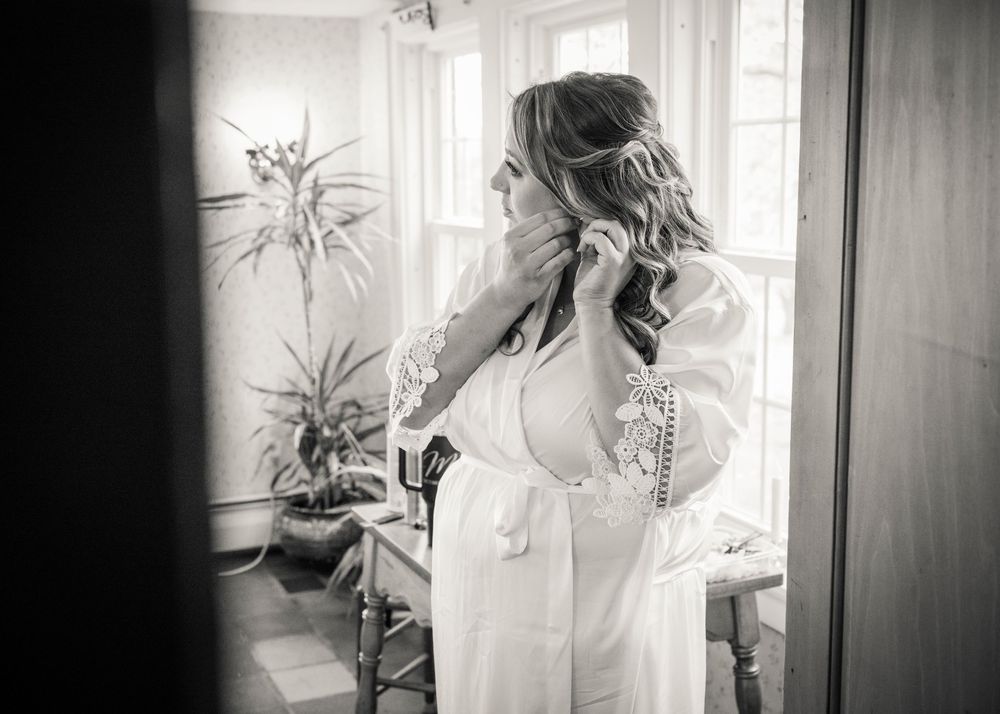 Black and white photo of a bride in a lace-trimmed robe putting on an earring by a sunlit window with a potted plant nearby.