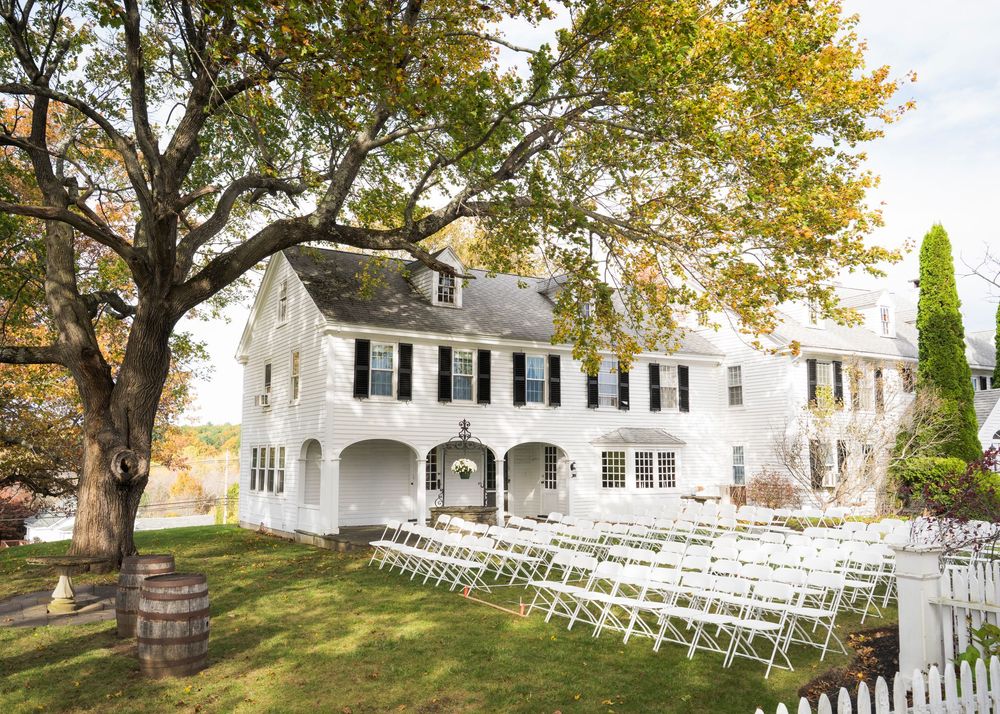 White colonial inn with black shutters and arched porch, with rows of white chairs set up on the lawn for an outdoor wedding ceremony.