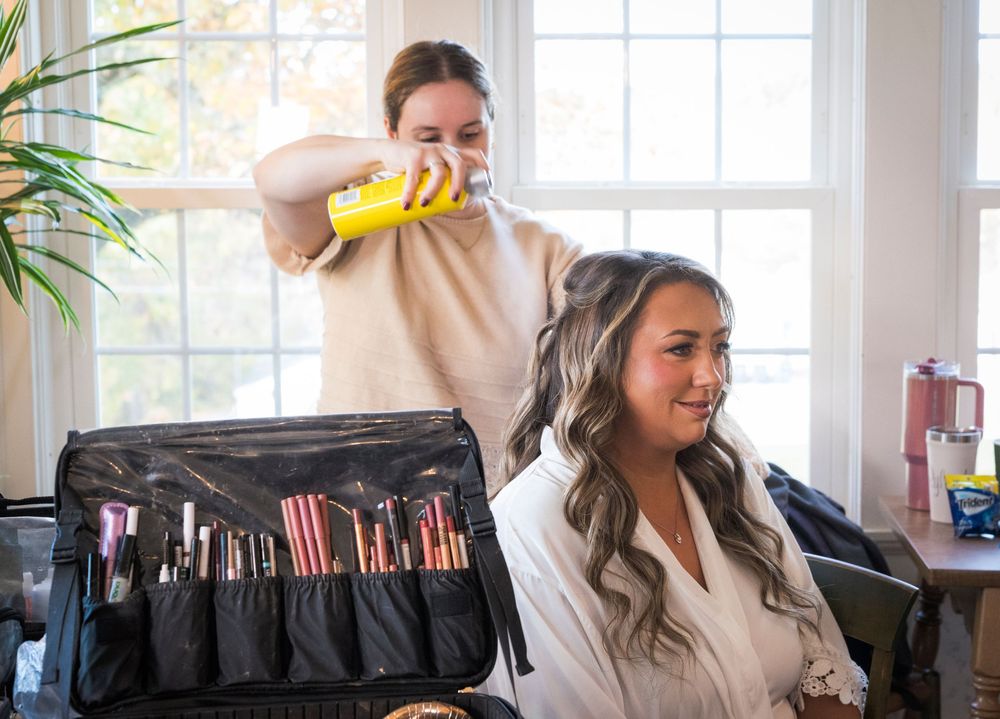 A stylist sprays hair product onto a smiling woman's long wavy curls beside an open makeup brush case in a bright, window-lit room.