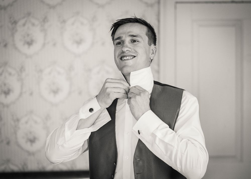 Smiling groom in a vest and white dress shirt adjusts his collar while getting dressed in a room with vintage floral wallpaper.