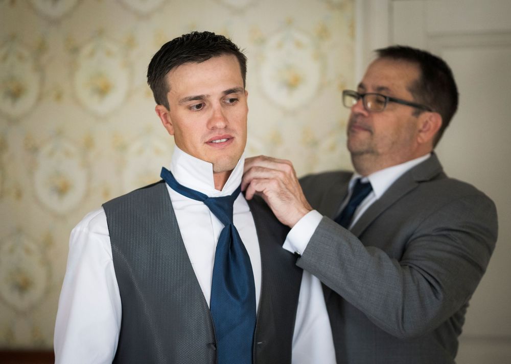 A groomsman in a gray suit helps the groom in a vest and white shirt adjust his blue necktie before the wedding ceremony.