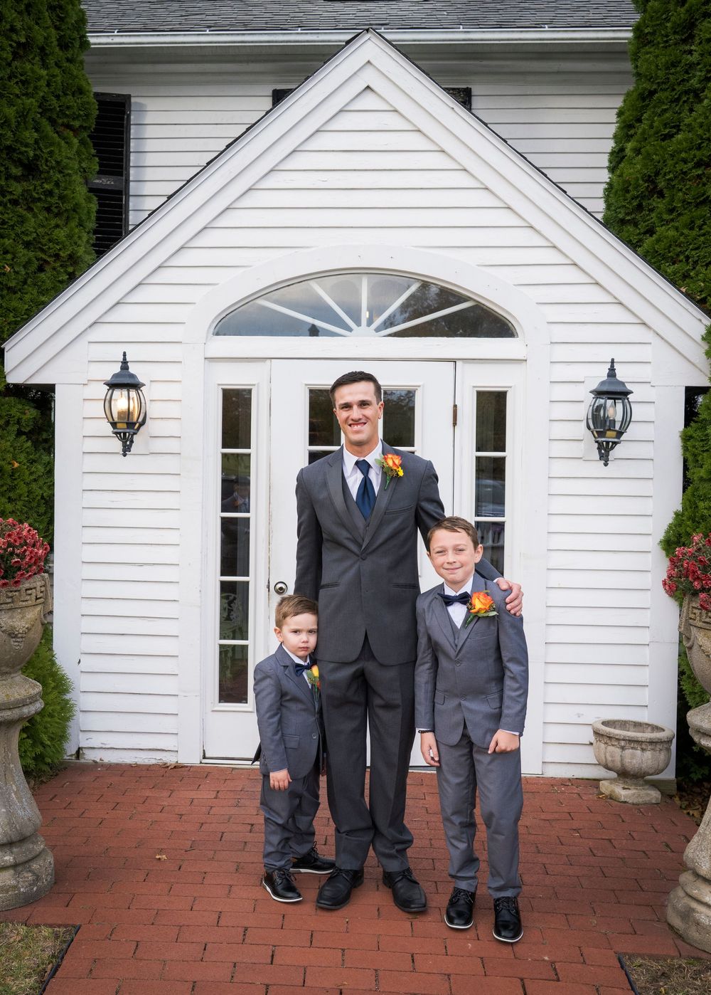 Groom in a gray suit and boutonniere stands with two young boys in matching suits on a brick patio outside a white colonial building.