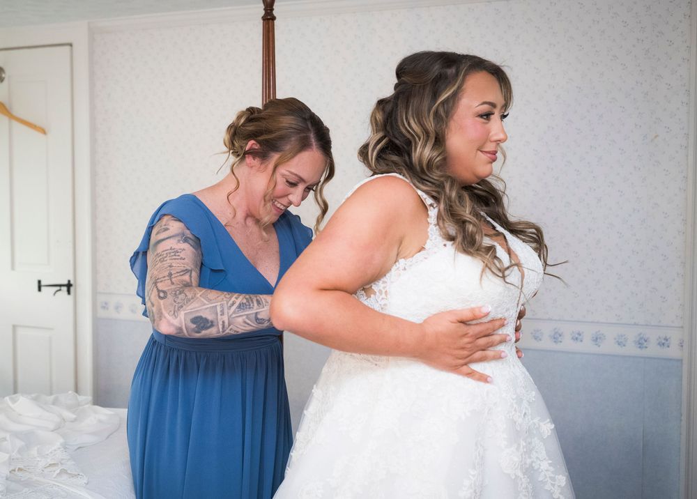 Bridesmaid in a blue dress with arm tattoos helps a bride in a white lace gown get ready in a bright bedroom.