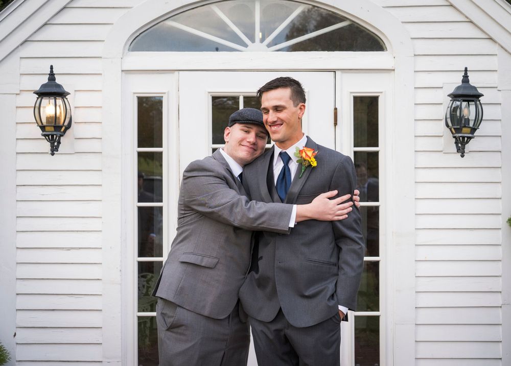 Two grooms in gray suits embrace and smile in front of a white chapel door flanked by black lanterns on their wedding day.