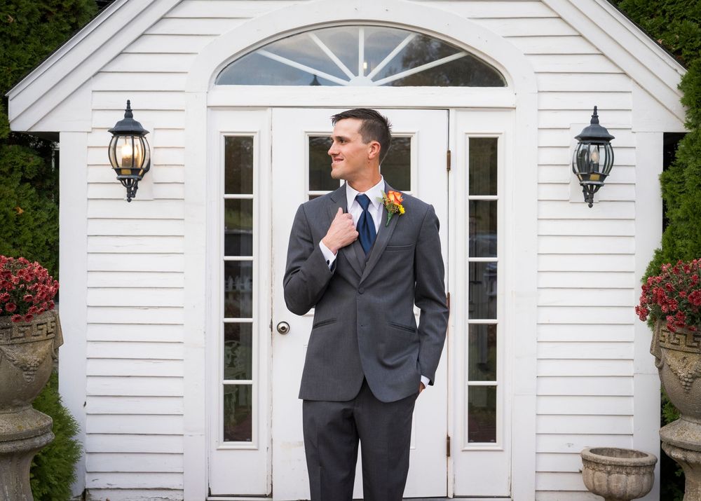 Groom in a gray suit and navy tie adjusts his jacket in front of a white chapel entrance flanked by lanterns and flowers.