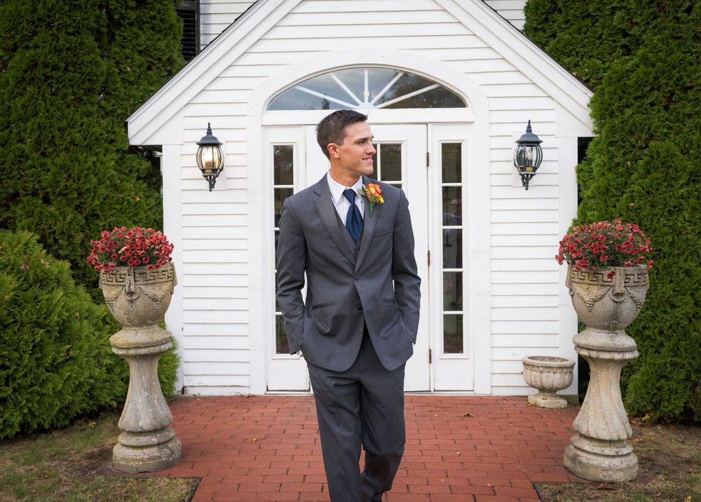 Groom in a gray suit and blue tie with a boutonniere stands on a brick path before a white chapel flanked by stone urns of red flowers.