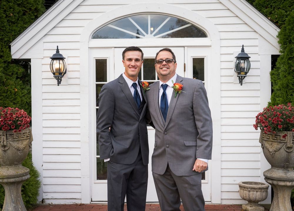 Two smiling groomsmen in gray suits and navy ties pose with boutonnieres in front of a white building with flower urns.