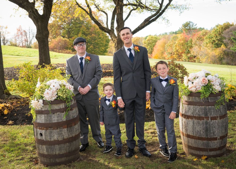 Groom poses with two young ring bearers in suits and bow ties and an older man among barrels of flowers and autumn trees.
