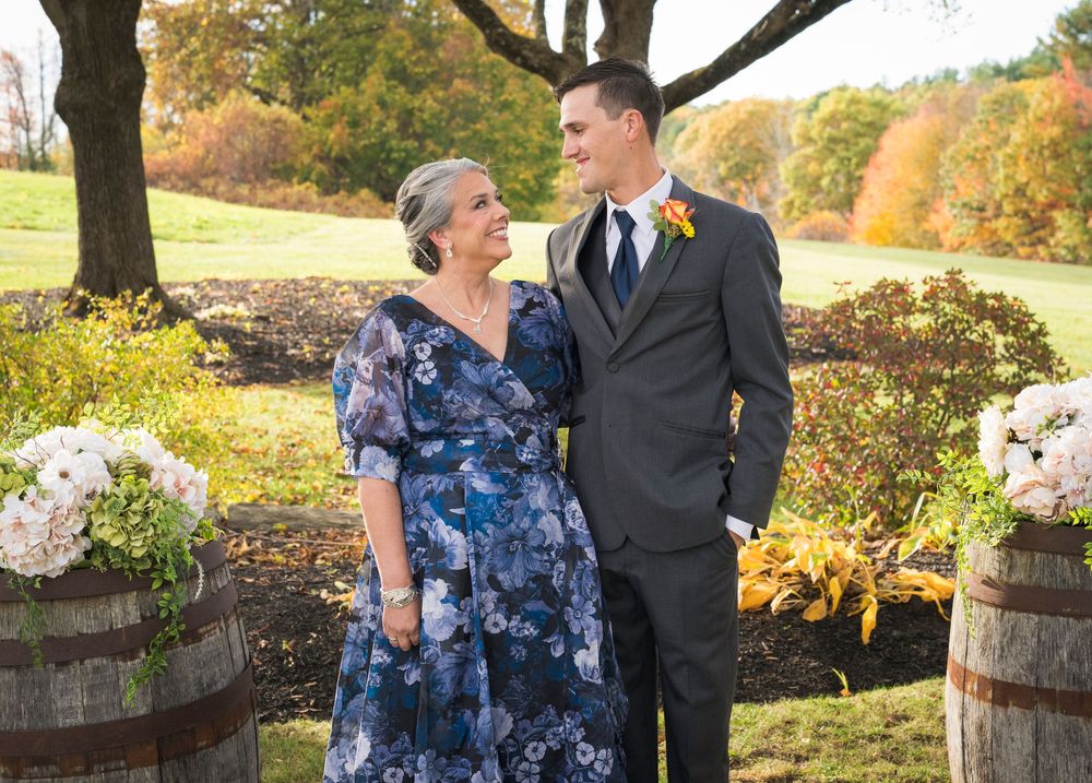Smiling woman in a blue floral dress stands beside a man in a gray suit with an orange boutonniere at an outdoor autumn wedding.