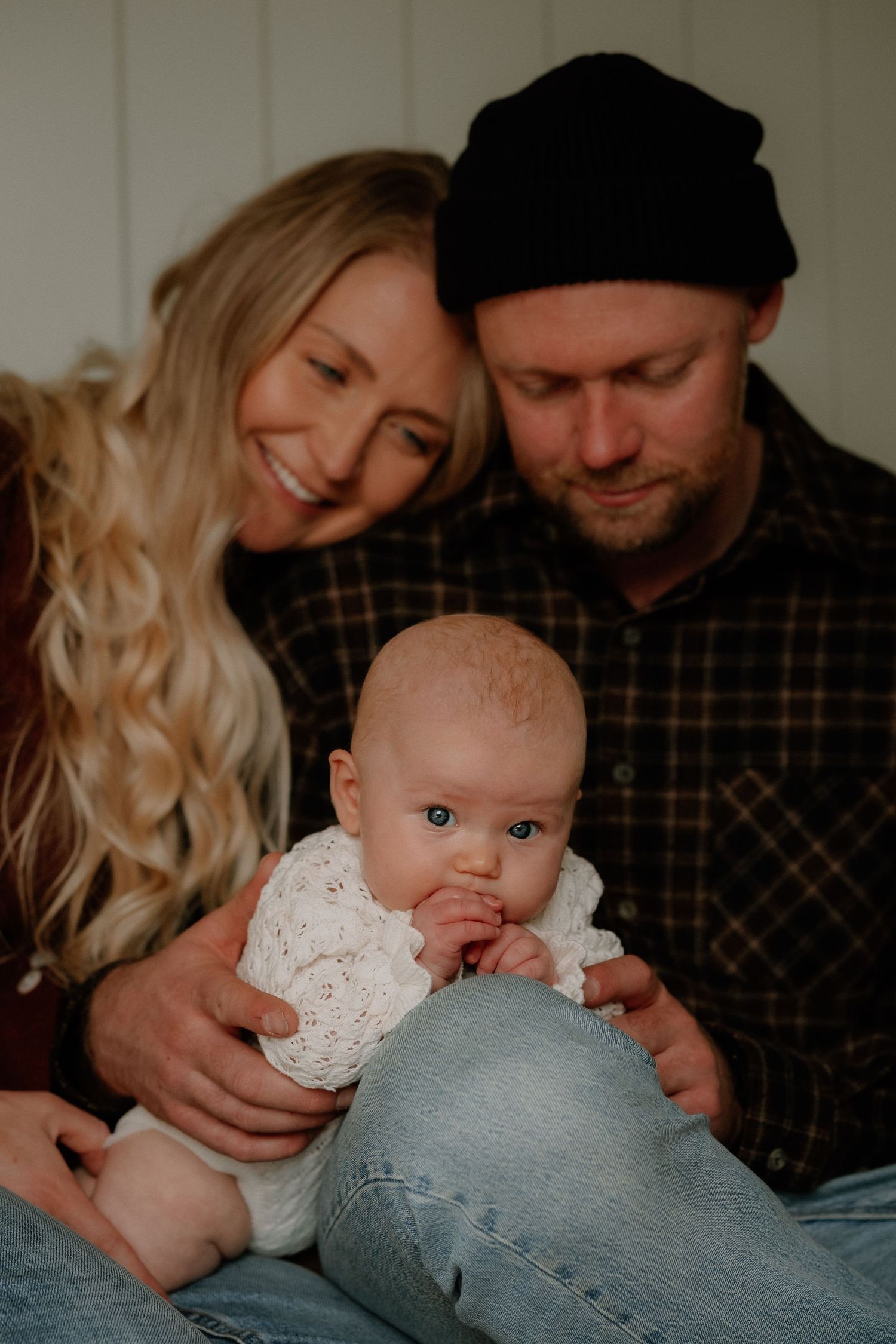 Happy young family with blonde mother, bearded father in black beanie, and baby in white knit outfit posing together.