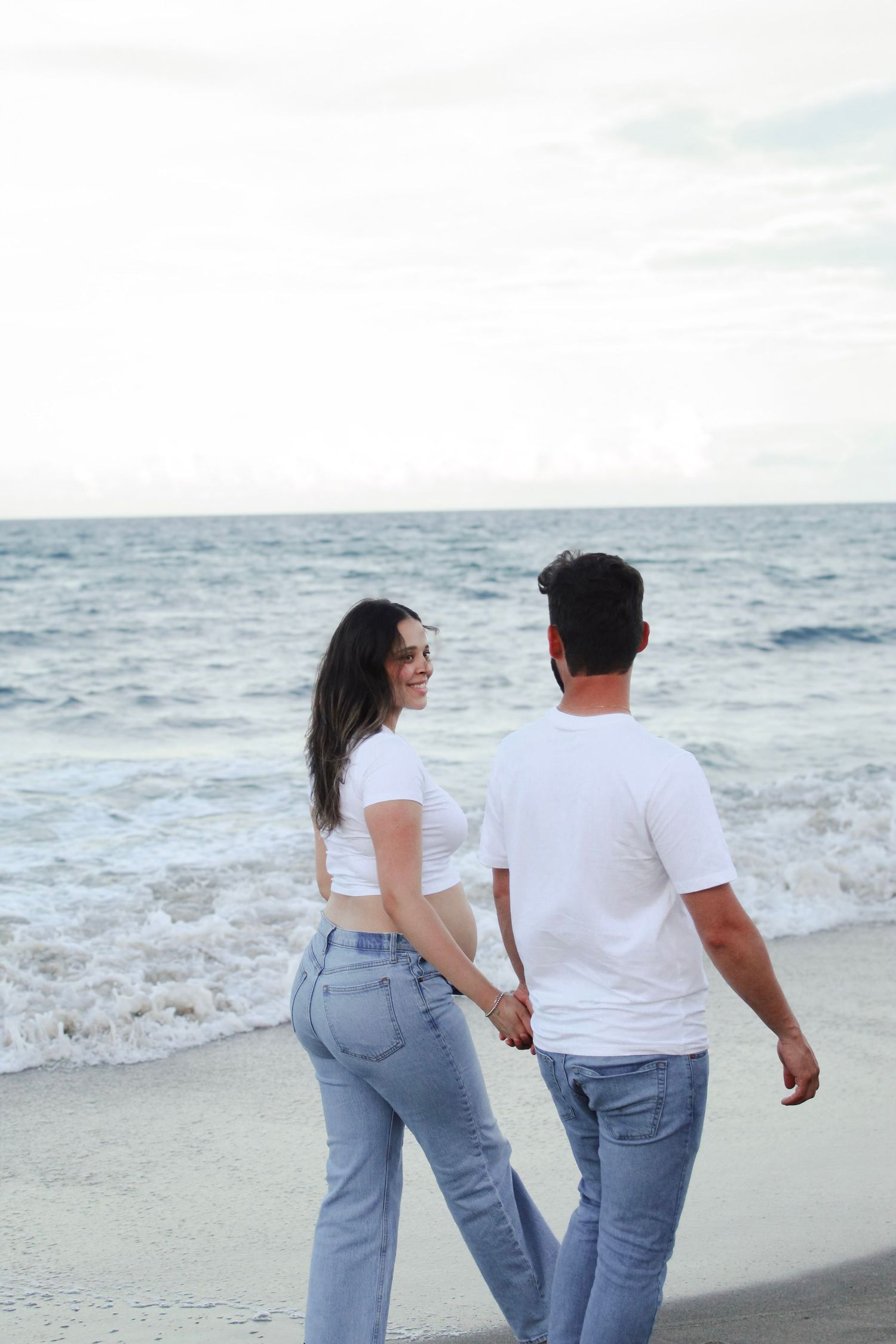Couple holding hands walking along sandy beach shore with ocean waves behind them.