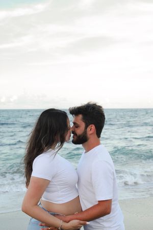 Bearded man and pregnant woman in white tops sharing a kiss on a beach.
